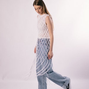 May include: A woman wearing a white crochet top with a net-like design and blue jeans. The top is long and has a fringe detail. The woman is standing on a white background.