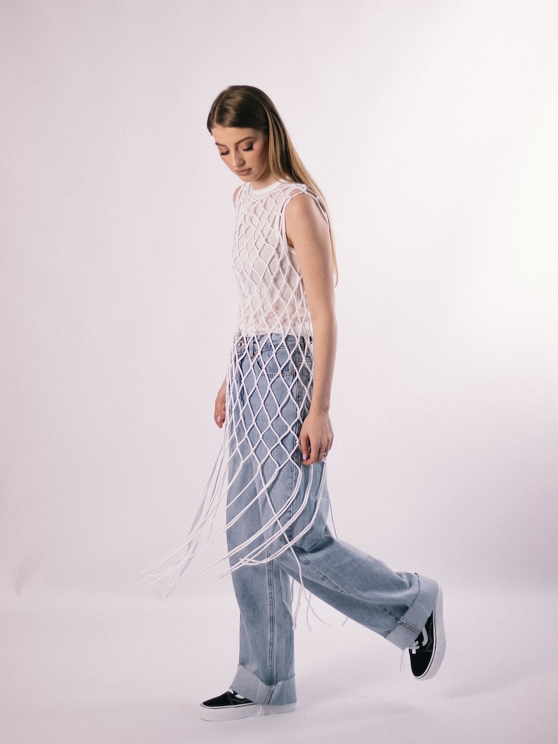 May include: A woman wearing a white crochet top with a net-like design and blue jeans. The top is long and has a fringe detail. The woman is standing on a white background.