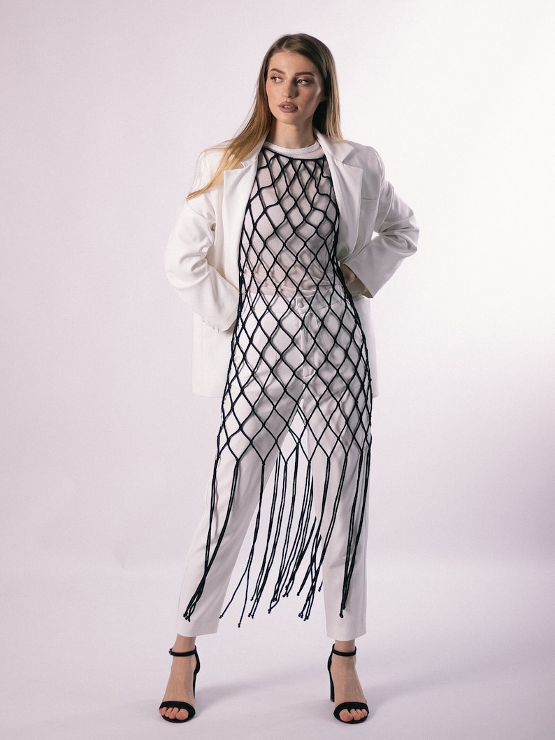 May include: A white blazer with a long, black, fringed mesh top and white pants. The model is wearing black heels.
