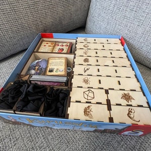 Puede incluir: Un inserto de madera para organizar el juego de mesa "Furioso: The Guild of Merchants". El inserto tiene compartimentos para cartas, fichas y otros componentes del juego. El inserto está hecho de madera marrón claro y tiene un borde azul y rojo.
