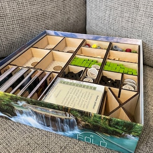Puede incluir: Un organizador de juegos de madera con compartimentos para guardar piezas de juego, cartas y fichas. El organizador tiene un diseño de cielo azul y blanco en la parte superior y una imagen de cascada en la parte inferior. El texto "FOLD" está impreso en la parte inferior del organizador.