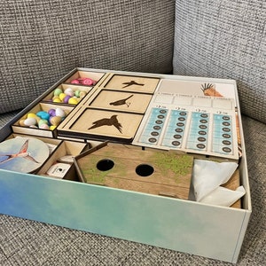 May include: A wooden game box with a blue and green gradient design. The box is open and contains various game components, including wooden bird figurines, a birdhouse, a score sheet, and a bag of colorful eggs.
