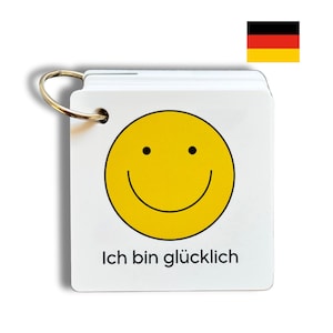 May include: A white card with a yellow smiley face and the text "Ich bin glücklich" in black. The card is attached to a silver ring.
