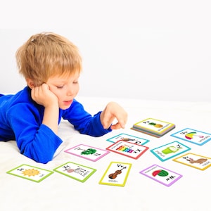 May include: A young child is playing with colorful alphabet flash cards featuring various images, including a sun, zebra, tree, xylophone, violin, watermelon, yogurt, pineapple, umbrella, and rabbit. The cards are laid out on a white surface.