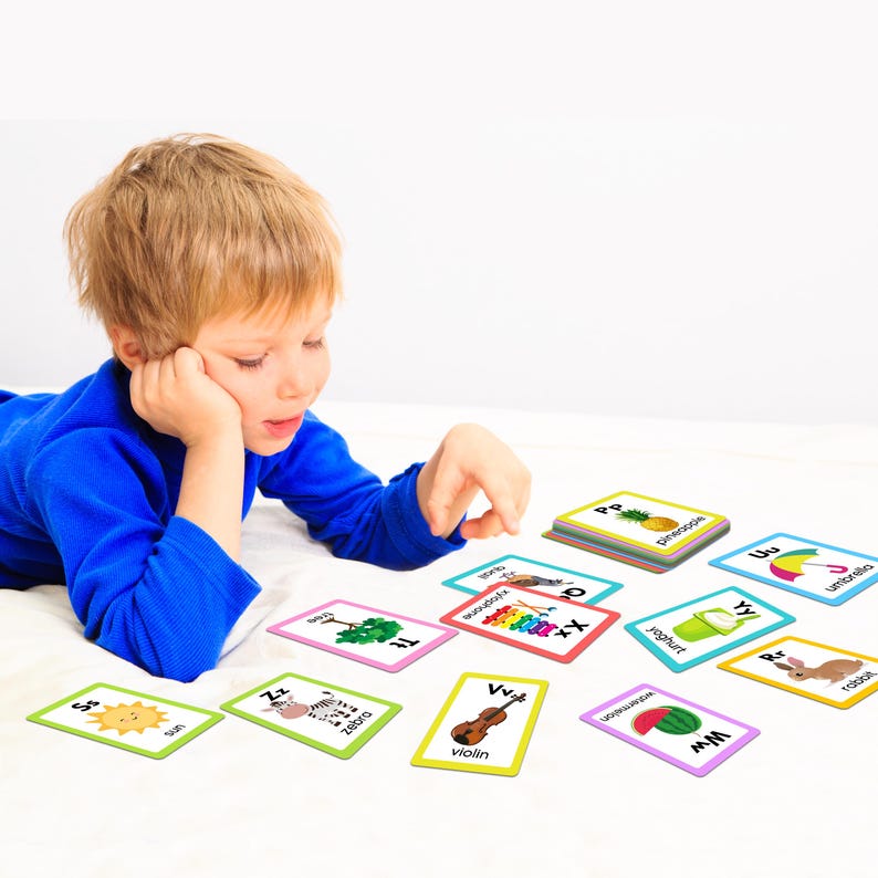 May include: A young child is playing with colorful alphabet flash cards featuring various images, including a sun, zebra, tree, xylophone, violin, watermelon, yogurt, pineapple, umbrella, and rabbit. The cards are laid out on a white surface.