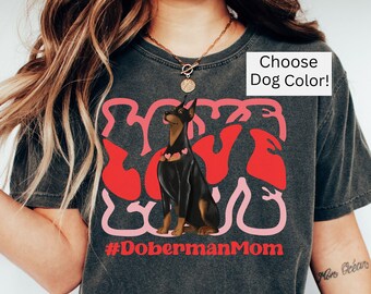 May include: A dark grey t-shirt with a pink and red graphic of the word "love" with a Doberman Pinscher dog sitting in the middle. The text "#DobermanMom" is below the graphic. The text "Choose Dog Color!" is in the top right corner of the image.
