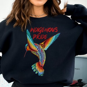 以下が含まれることがあります： 赤い文字で「Indigenous Pride」と書かれた、カラフルなハチドリのグラフィックがプリントされた黒いスウェットシャツです。