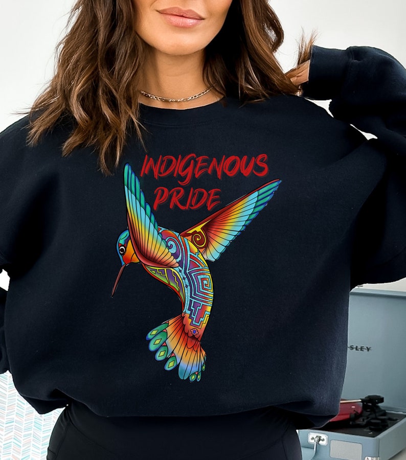以下が含まれることがあります： 赤い文字で「Indigenous Pride」と書かれた、カラフルなハチドリのグラフィックがプリントされた黒いスウェットシャツです。