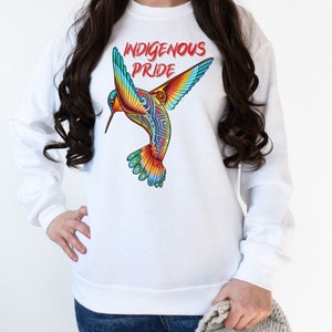 以下が含まれることがあります： 前面にカラフルなハチドリのグラフィックと「Indigenous Pride」というテキストがプリントされた白いスウェットシャツです。