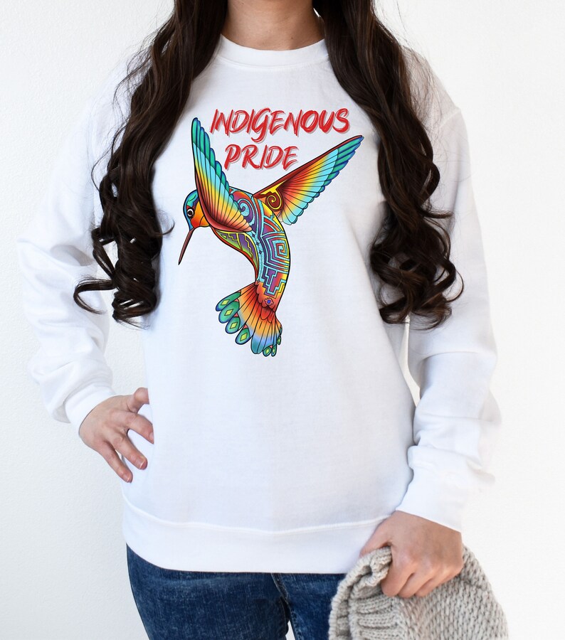 以下が含まれることがあります： 前面にカラフルなハチドリのグラフィックと「Indigenous Pride」というテキストがプリントされた白いスウェットシャツです。