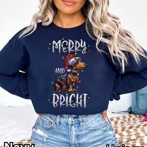 K&ouml;nnte beinhalten: Marineblauer Sweatshirt mit einer wei&szlig;en Grafik eines Dackels, der einen Weihnachtsmann-Hut tr&auml;gt und von Weihnachtslichtern umgeben ist. Der Text "Merry and Bright" ist auf dem Sweatshirt aufgedruckt.