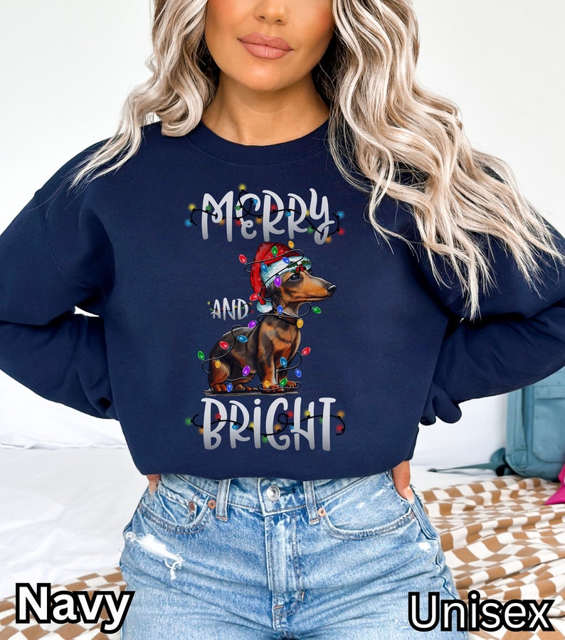 K&ouml;nnte beinhalten: Marineblauer Sweatshirt mit einer wei&szlig;en Grafik eines Dackels, der einen Weihnachtsmann-Hut tr&auml;gt und von Weihnachtslichtern umgeben ist. Der Text "Merry and Bright" ist auf dem Sweatshirt aufgedruckt.