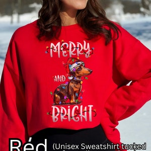 K&ouml;nnte beinhalten: Roter Unisex-Sweatshirt mit einem weihnachtlichen Design, das einen Dackel zeigt, der einen Weihnachtsmann-Hut tr&auml;gt und von bunten Lichtern umgeben ist. Der Text "Merry and Bright" ist auf dem Sweatshirt aufgedruckt.