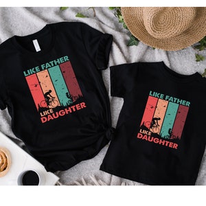 May include: Two black t-shirts with a vintage-style graphic that says "Like Father Like Daughter" and features a father and daughter riding bicycles. The graphic has a red, orange, yellow, green, and blue color scheme.