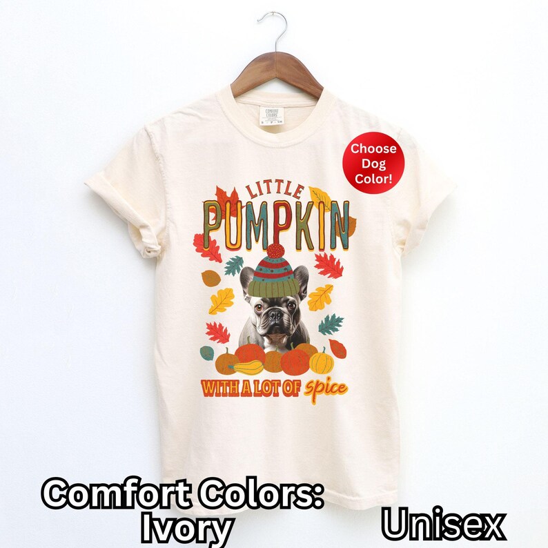 May include: A cream-colored t-shirt with a graphic of a dog wearing a hat and surrounded by pumpkins and fall leaves. The text "LITTLE PUMPKIN WITH A LOT OF spice" is printed on the shirt. The shirt is available in a variety of colors. Comfort Colors: Ivory. Unisex.