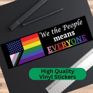 May include: A black and white sticker with a rainbow pride flag, an American flag, and a transgender pride flag. The sticker says "We the People means EVERYONE" in white text.