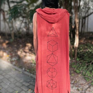 Può includere: Un capo senza maniche con cappuccio, color corallo, con forme geometriche nere stampate sul retro. La persona indossa un berretto marrone. Il capo ha uno spacco laterale. Lo sfondo è sfocato con vegetazione.