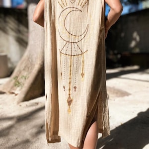 Könnte beinhalten: Beige ärmelloser Kapuzenponcho mit einem Himmelsdesign. Die Rückseite des Ponchos zeigt ein Halbmond- und Sonnenaufgangsdesign in Braun. Der Poncho hat an jeder Seite einen Schlitz und ist knielang.