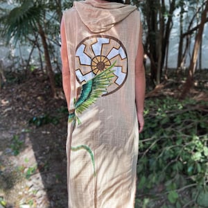 Könnte beinhalten: Beigefarbener ärmelloser Kapuzenmantel mit großem kreisförmigen Design auf dem Rücken, das ein sonnenähnliches Symbol und eine grün-blaue Feder zeigt. Der Mantel ist aus leichtem Stoff gefertigt und hat eine lange, fließende Silhouette.