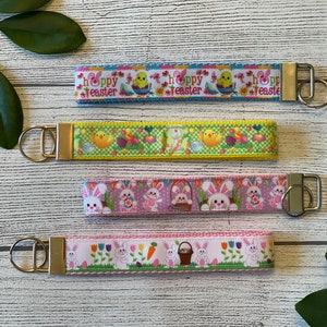 May include: Four colorful Easter-themed keychains with different designs. The keychains feature bunnies, chicks, eggs, and flowers. The keychains are attached to gold-colored key rings.
