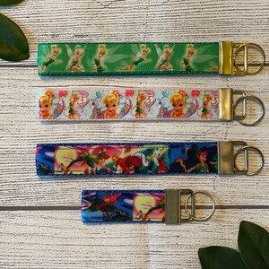 May include: Four different keychains featuring Tinkerbell and Peter Pan characters. The keychains are made of fabric with colorful images of the characters. The keychains are attached to gold and silver key rings.