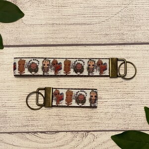 May include: Two keychains with a white ribbon featuring cartoon characters and bronze-colored hardware. The larger keychain has a repeating pattern of characters. The smaller keychain has a similar design. The keychains are on a light wood surface.