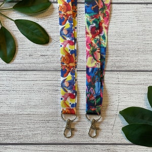 May include: Two colorful lanyards with cartoon character designs. The lanyard on the left features Winnie the Pooh characters, while the lanyard on the right features characters from Peter Pan. Both lanyards have silver swivel clips.