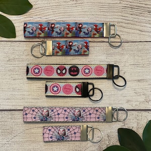 May include: Five different keychains with various superhero designs. The keychains feature a white background with colorful superhero images, including Spider-Man, Captain America, Superman, and Batman. The keychains are attached to silver key rings.
