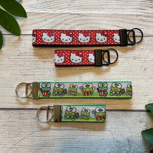 May include: Four keychains with different cartoon characters. The top two keychains feature a red and white Hello Kitty design. The bottom two keychains feature a green and white Keropi design.