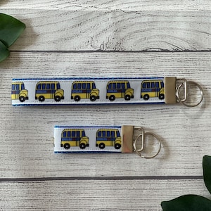 May include: Two yellow school bus keychains with blue trim and white background. The larger keychain has five buses and the smaller keychain has two buses. Both keychains have silver key rings.