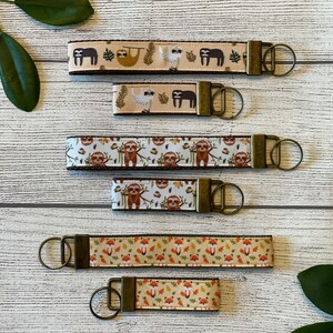 May include: Five keychains with different sloth and fox designs. The keychains are made of brown and beige ribbon with a gold metal key ring. The keychains are arranged in a row on a white wooden surface.