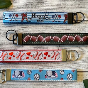 May include: Four different sports-themed keychains with colorful ribbon straps and metal key rings. The keychains feature designs of hockey, football, baseball, and volleyball.