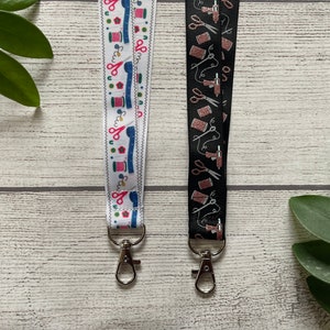 May include: Two colorful fabric lanyards with silver keychains. One lanyard is white with blue, red, and pink sewing themed designs. The other lanyard is black with rose gold sewing themed designs.