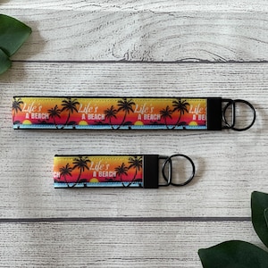 May include: Two black keychains with a colorful sunset and palm tree design. The keychains feature the text "Life's a Beach".