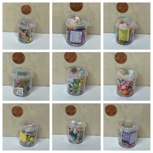 May include: Nine small, clear plastic containers filled with miniature objects. Each container has a clear lid and a small handle. The objects inside vary, including dice, playing cards, and small paper labels. A penny is placed near each container.