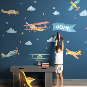 May include: A child's room decorated with a blue wall and colorful airplane wall decals. The decals include clouds, stars, and airplanes. The name "JOHN" is written on a banner attached to one of the airplanes. A child is holding a toy airplane in the room.