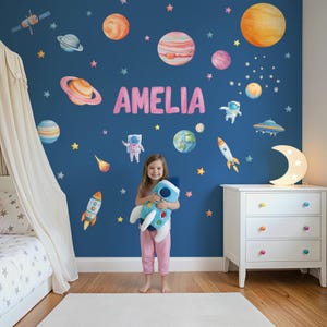 Può includere: Camera da letto per bambini con una parete blu decorata con adesivi a tema spaziale, tra cui pianeti, stelle e razzi. Il nome "AMELIA" è visualizzato in lettere rosa. Una bambina tiene in mano un peluche a forma di razzo blu davanti a un comò bianco.