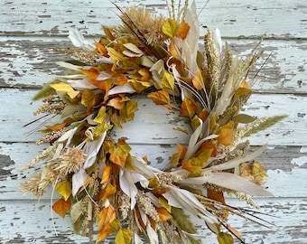 May include: A fall-themed wreath with dried leaves, grasses, and flowers in shades of beige, brown, and yellow. The wreath is made of natural materials and has a rustic look.