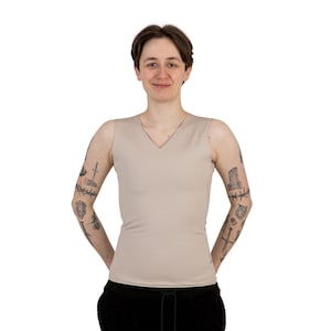 May include: A person wearing a sleeveless, light beige v-neck top. The person has tattoos on both arms.