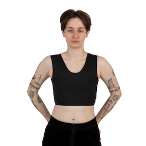 May include: A black sleeveless crop top with a scoop neck. The model is wearing the top and has tattoos on their arms.