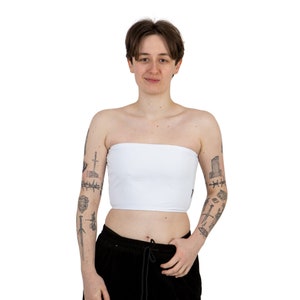 May include: A white strapless tube top with a smooth, matte finish. The top is worn by a person with short, dark hair and tattoos on their arms.