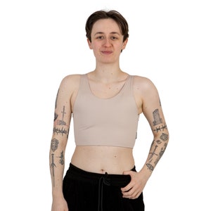 Puede incluir: Una persona lleva una camiseta de tirantes beige con cuello redondo. La camiseta de tirantes es corta y tiene un corte holgado. La persona tiene tatuajes en los brazos.