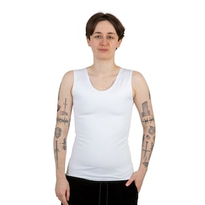 Puede incluir: Una camiseta blanca sin mangas con cuello redondo. La persona que lleva la camiseta tiene varios tatuajes en los brazos.
