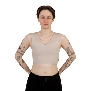 May include: A beige, V-neck chest binder. The binder is a cropped style, with a smooth, solid color. The person in the image is wearing black pants. The background is plain white.