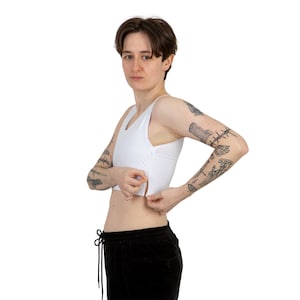 Puede incluir: Un top de compresión blanco, corto, con cierre de cremallera. El top lo lleva una persona con tatuajes en los brazos.