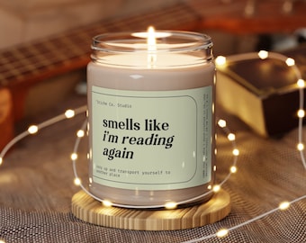 May include: A brown candle with a white label that reads "smells like i'm reading again" and "cozy up and transport yourself to another place". The candle is lit and sitting on a wooden coaster.