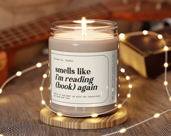 May include: A brown candle with a white label that reads "smells like I'm reading (book) again". The candle is sitting on a wooden coaster and surrounded by white string lights.