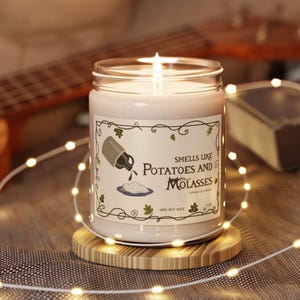 May include: A lit candle in a glass jar with a label that reads "Smells Like Potatoes and Molasses." The candle sits on a wooden coaster, surrounded by string lights. The candle is made of 100% soy wax.