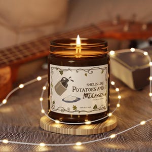 May include: A brown glass candle with a white label that reads "Smells like Potatoes and Molasses". The candle is lit and sitting on a wooden coaster.