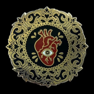 May include: Gold-toned enamel pin with a black background and a red anatomical heart with a white eye in the center. The heart is surrounded by a gold-toned ornate design.